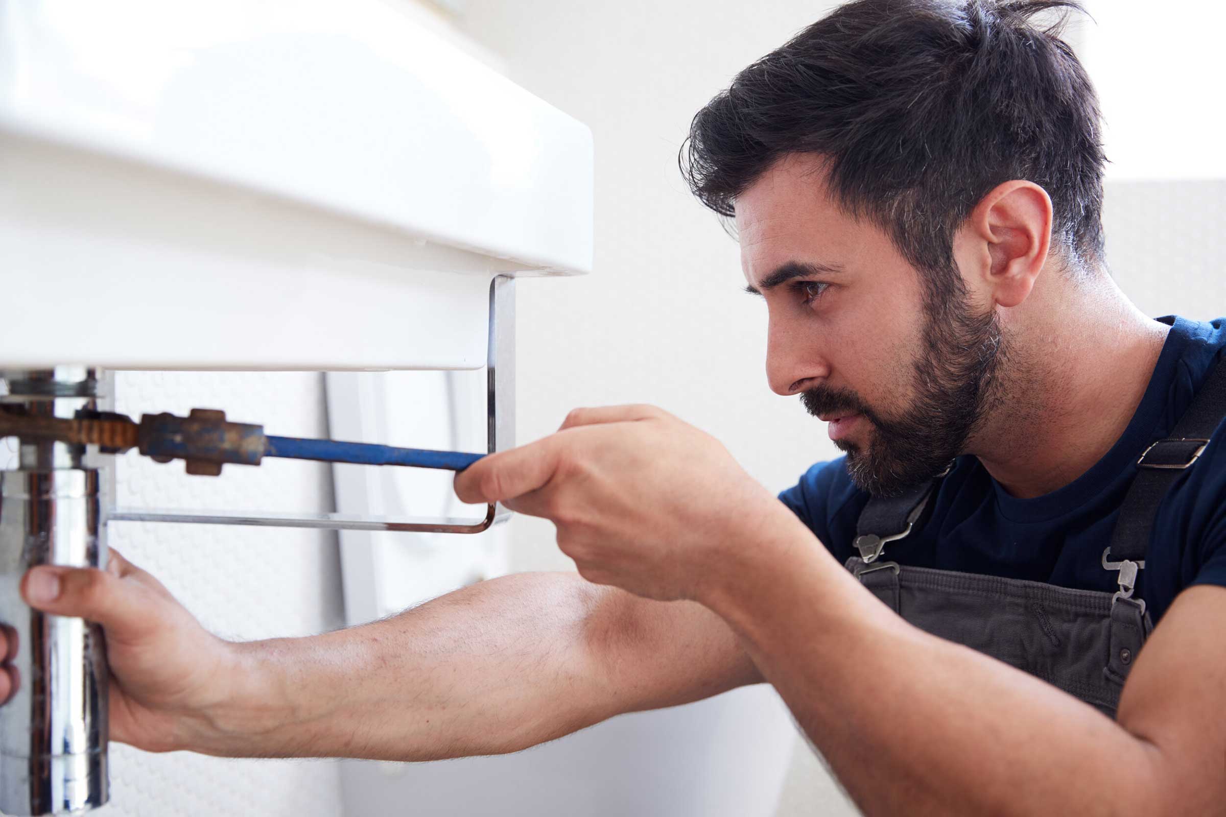 Male Plumber Using Wrench To Fix Leaking Sink In Home Bathroom