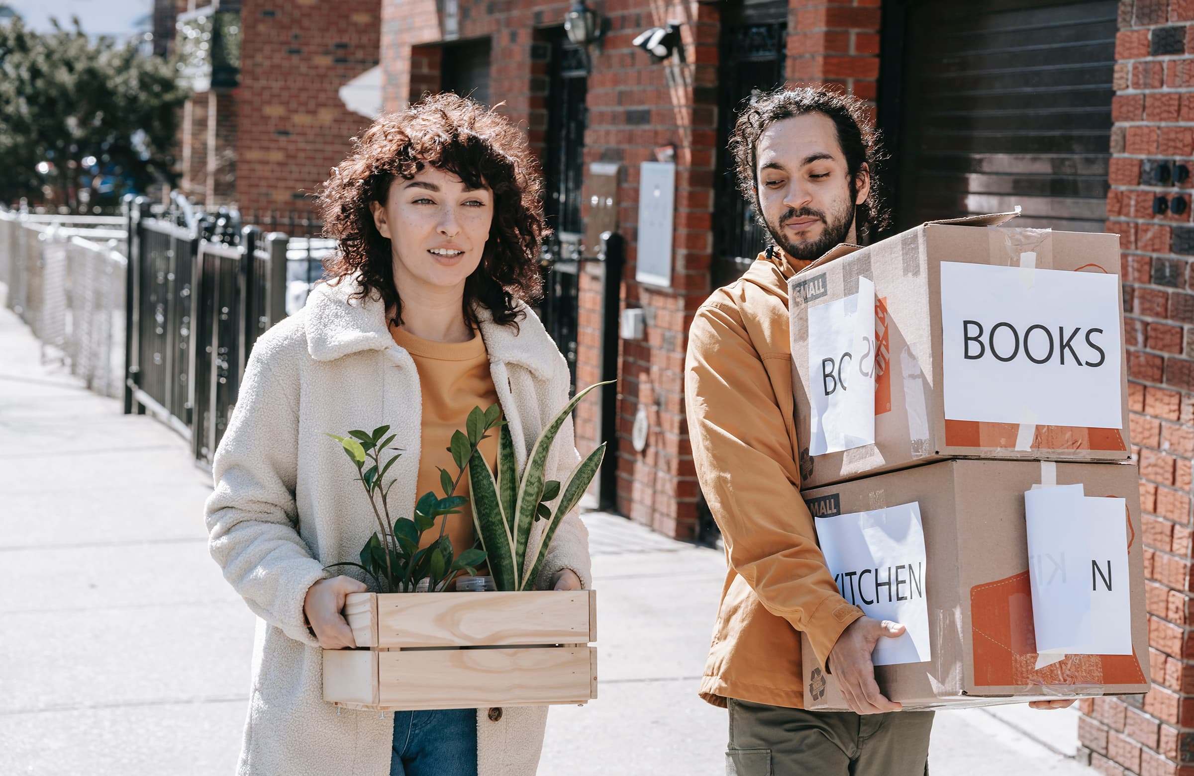 Couple Walking In The Street Carrying Plants And Boxes